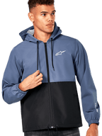 Modello uomo che indossa la giacca frangivento Alpinestars Speeder, design a blocchi di colore blu ardesia e nero, con cappuccio e cordini neri, logo bianco sul petto, giacca a vento casual ispirata al motorsport