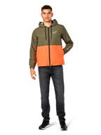 Giacca a vento Alpinestars Speeder, design color-block verde militare e arancione, giacca leggera con cappuccio, cordini neri e zip frontale, logo bianco su petto e manica, indossata con jeans scuri e sneaker nere, stile casual sportswear