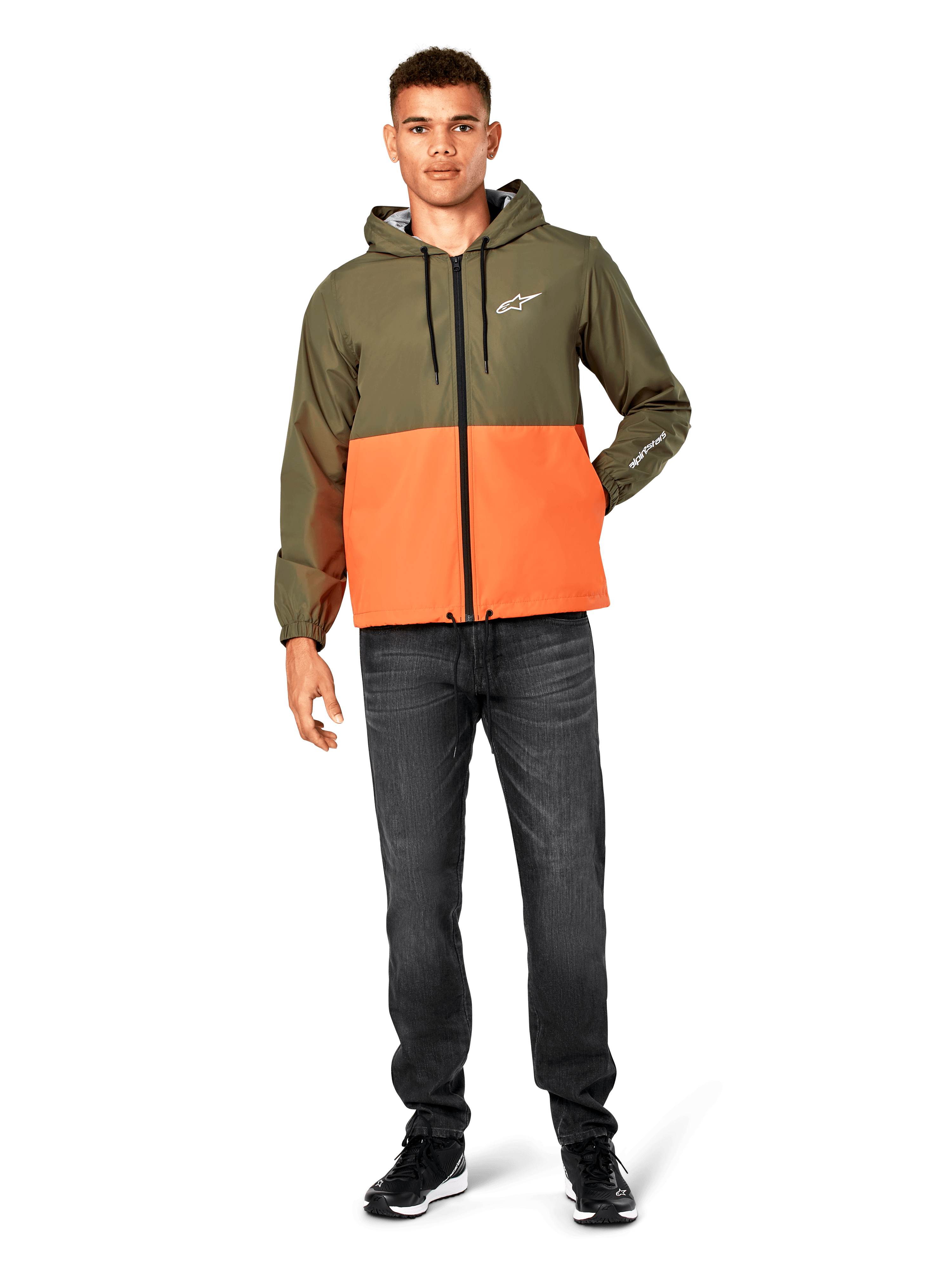 Giacca a vento Alpinestars Speeder, design color-block verde militare e arancione, giacca leggera con cappuccio, cordini neri e zip frontale, logo bianco su petto e manica, indossata con jeans scuri e sneaker nere, stile casual sportswear