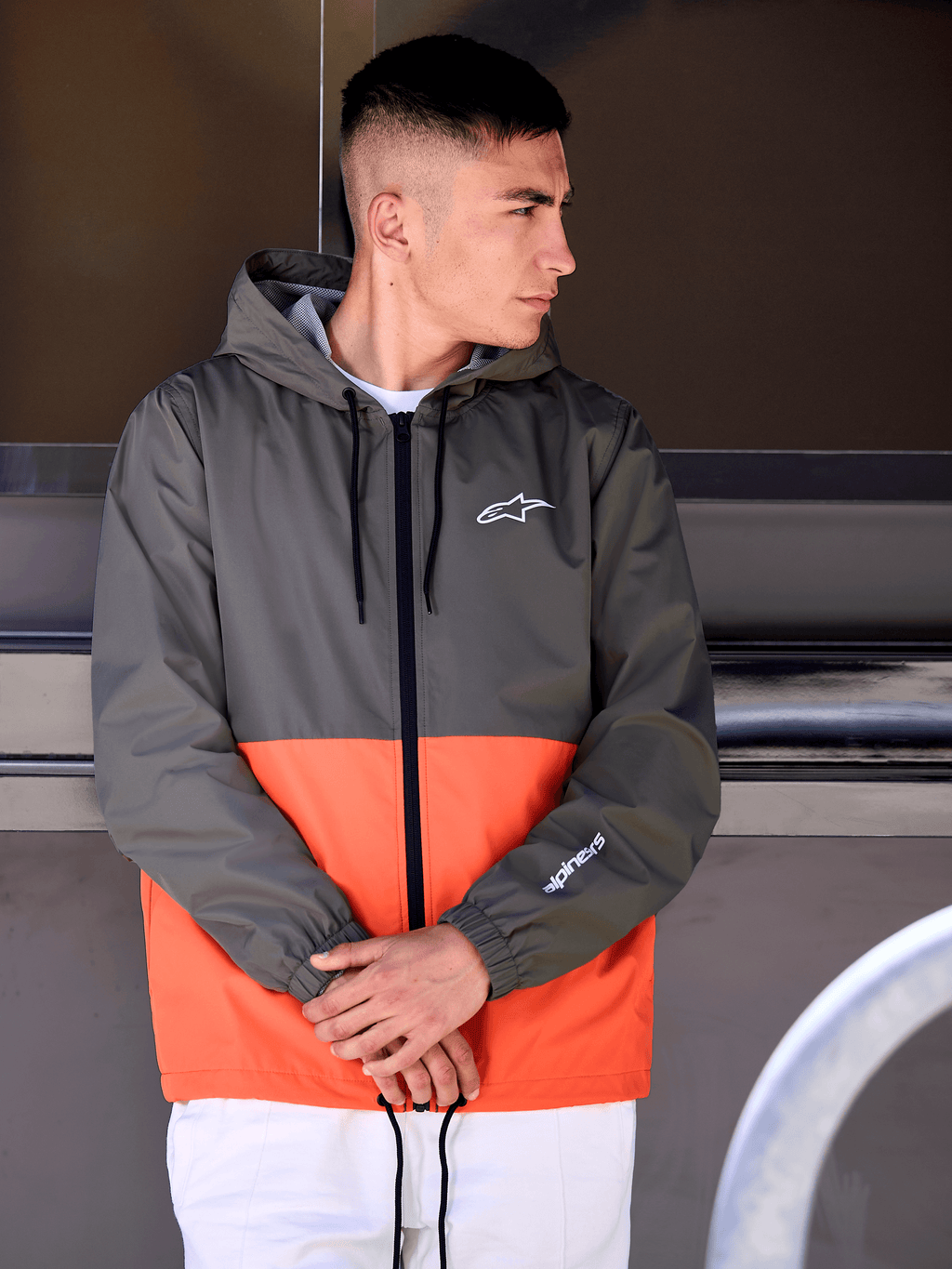 Uomo con giacca a vento Alpinestars Speeder, design a blocchi di colore arancione militare e verde scuro, giacca casual con cappuccio e loghi bianchi su petto e manica, abbinata a pantaloni bianchi per un look lifestyle ispirato al motorsport