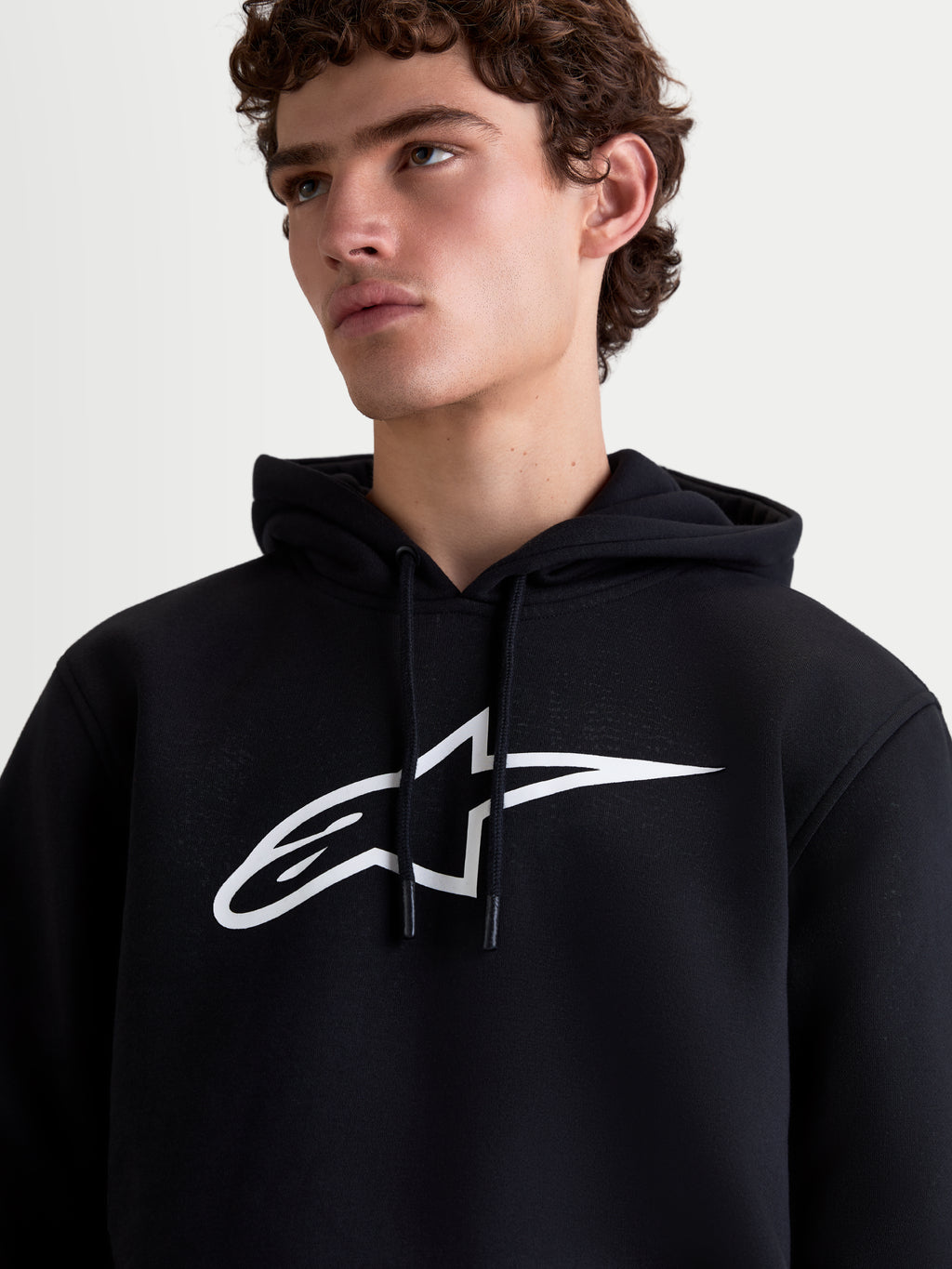 Alpinestars Felpa con cappuccio Ageless V3, felpa pullover da uomo in pile, nera e bianca, vista ravvicinata su modello con grande logo Astars bianco sul petto e cappuccio con cordino, stile casual motorsport.