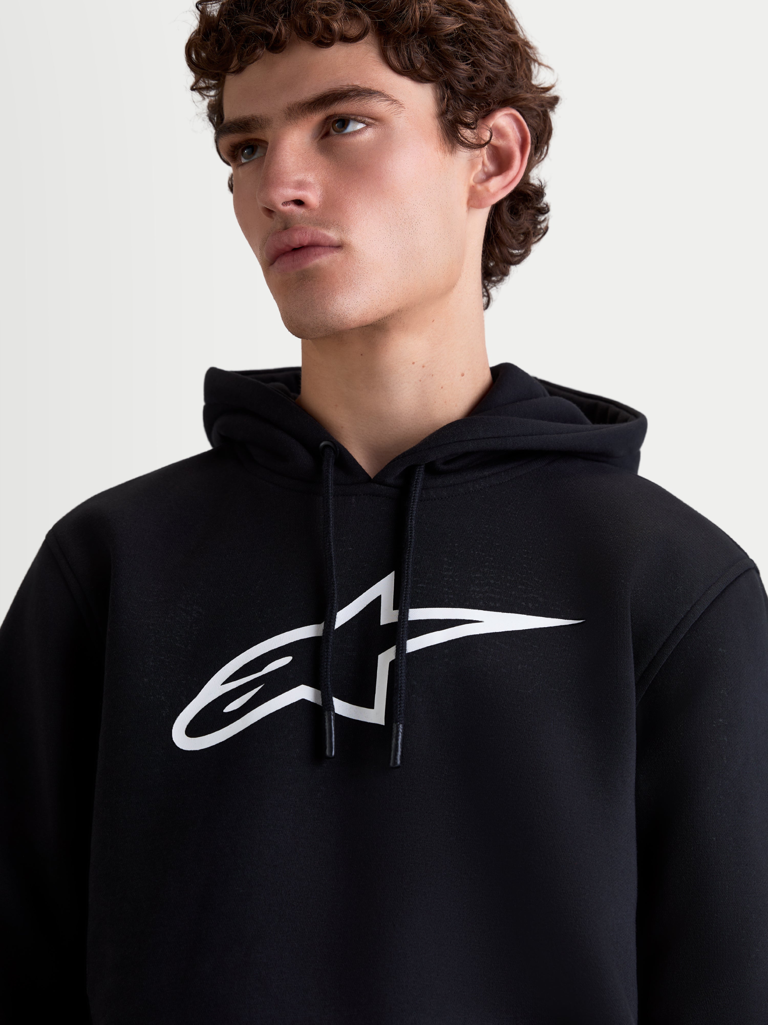 Alpinestars Felpa con cappuccio Ageless V3, felpa pullover da uomo in pile, nera e bianca, vista ravvicinata su modello con grande logo Astars bianco sul petto e cappuccio con cordino, stile casual motorsport.