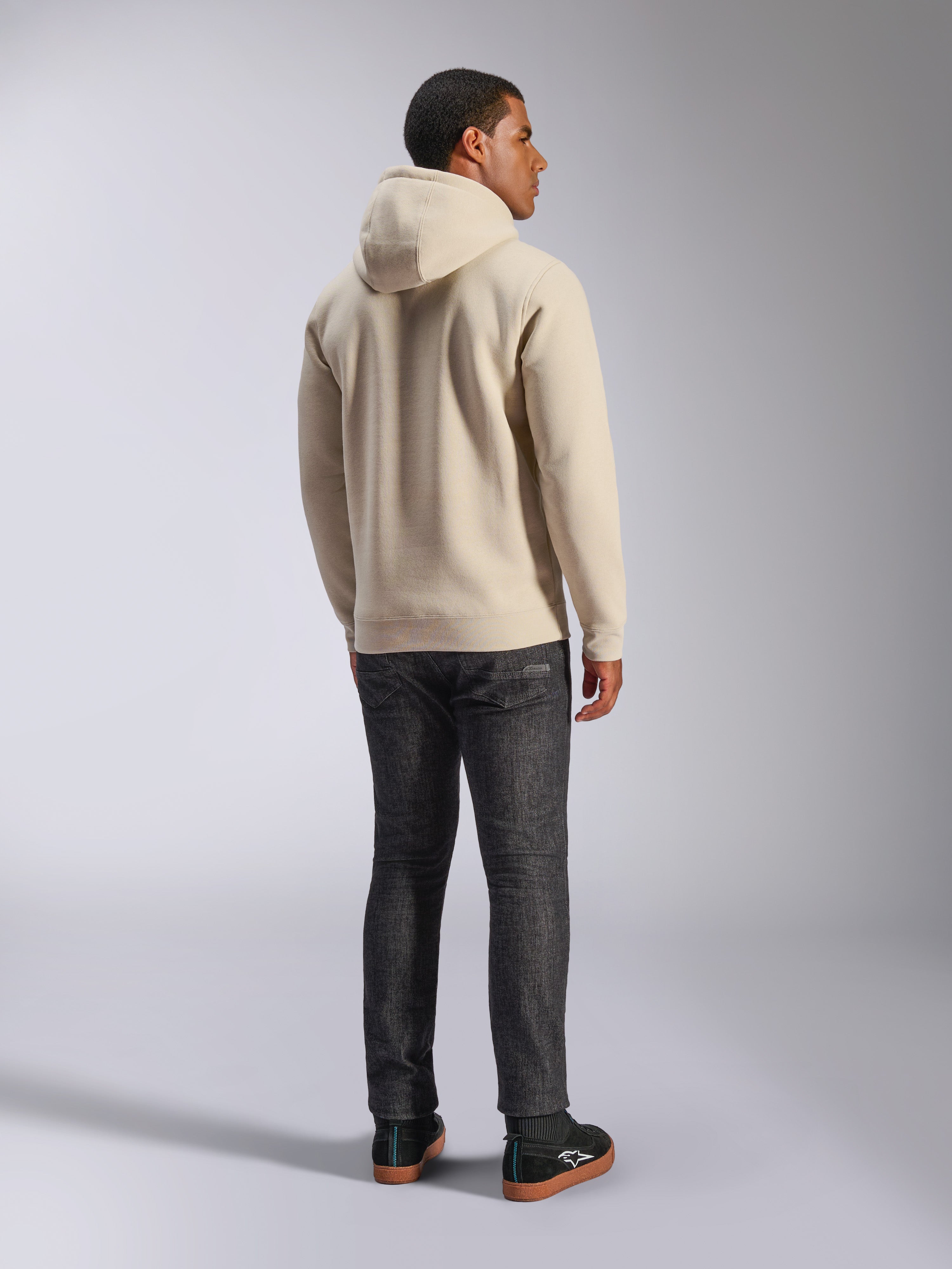 Alpinestars Ageless V3 Hoodie, felpa casual con cappuccio in pile bianco sporco vista dal retro, caratterizzata da polsini e orlo a costine, indossata con jeans scuri slim-fit e sneakers Alpinestars nere