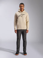 Alpinestars Ageless V3 Hoodie, abbigliamento sportivo casual, bianco sporco con logo nero ricamato, indossata da un uomo con pantaloni grigio scuro e sneakers nere, con cappuccio con coulisse e tasca a marsupio, design moderno ispirato al motorsport