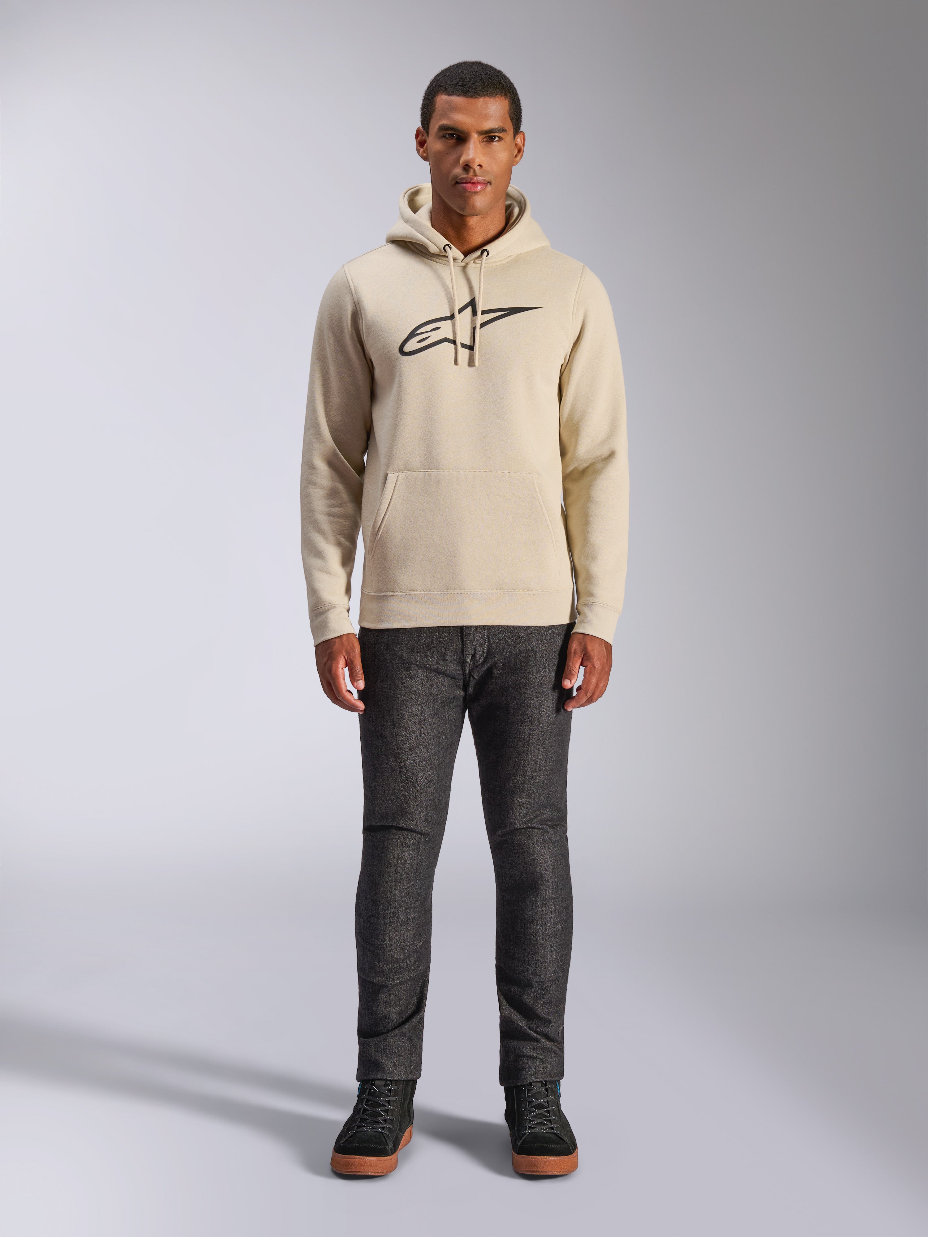 Alpinestars Ageless V3 Hoodie, abbigliamento sportivo casual, bianco sporco con logo nero ricamato, indossata da un uomo con pantaloni grigio scuro e sneakers nere, con cappuccio con coulisse e tasca a marsupio, design moderno ispirato al motorsport
