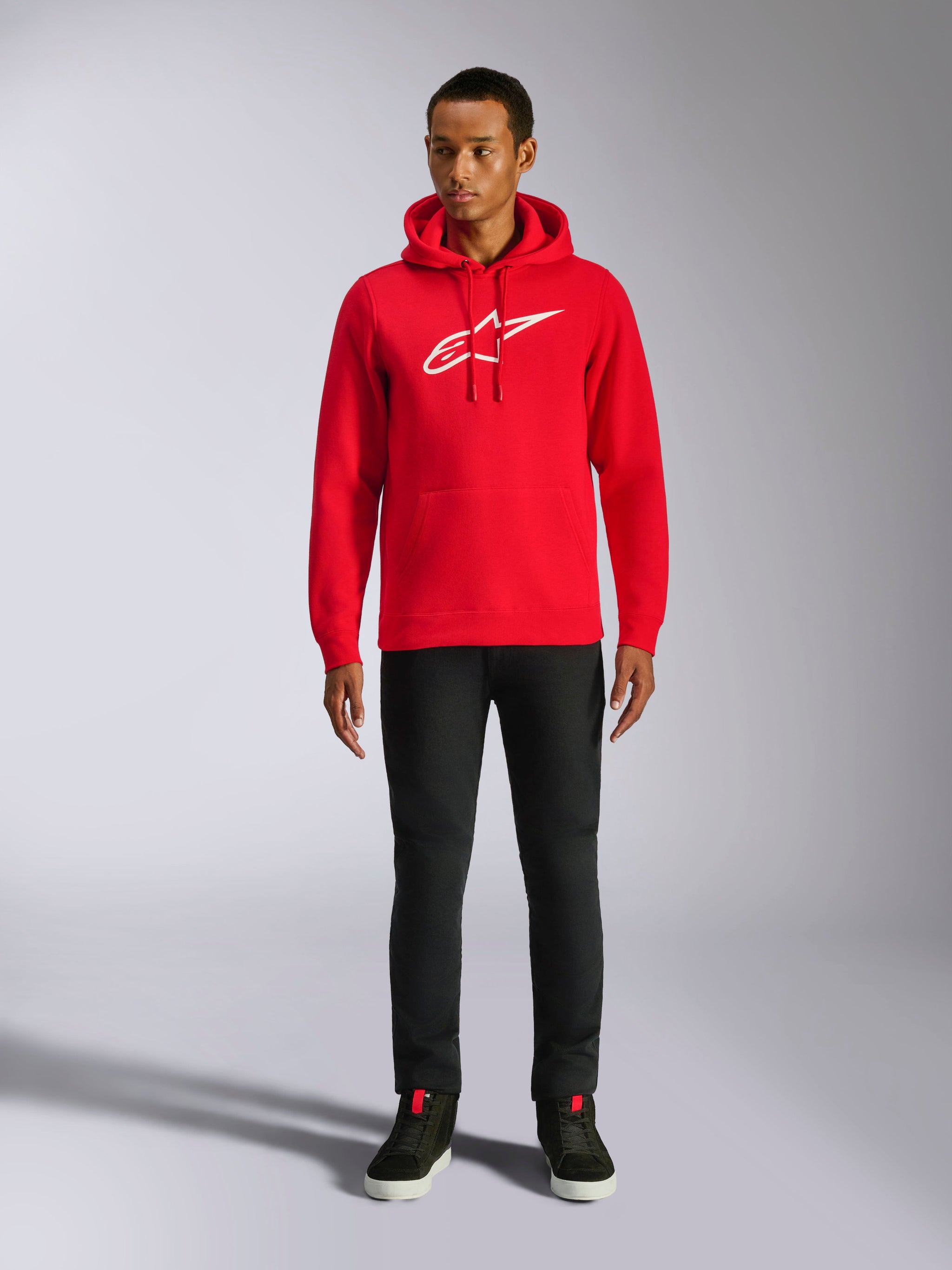 Felpa con cappuccio Alpinestars Ageless V3, streetwear casual, rossa con logo Alpinestars bianco ricamato, indossata da un modello con pantaloni neri, cappuccio con coulisse, tasca a marsupio anteriore, design moderno ispirato al motorsport
