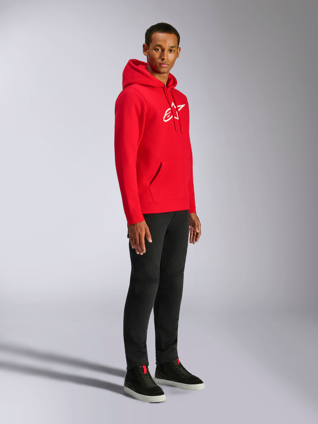 Alpinestars Ageless V3 Hoodie, felpa con cappuccio rossa con logo ricamato bianco, felpa sportiva casual indossata da un uomo con pantaloni neri e sneakers nere, con cappuccio con cordoncino e polsini a costine
