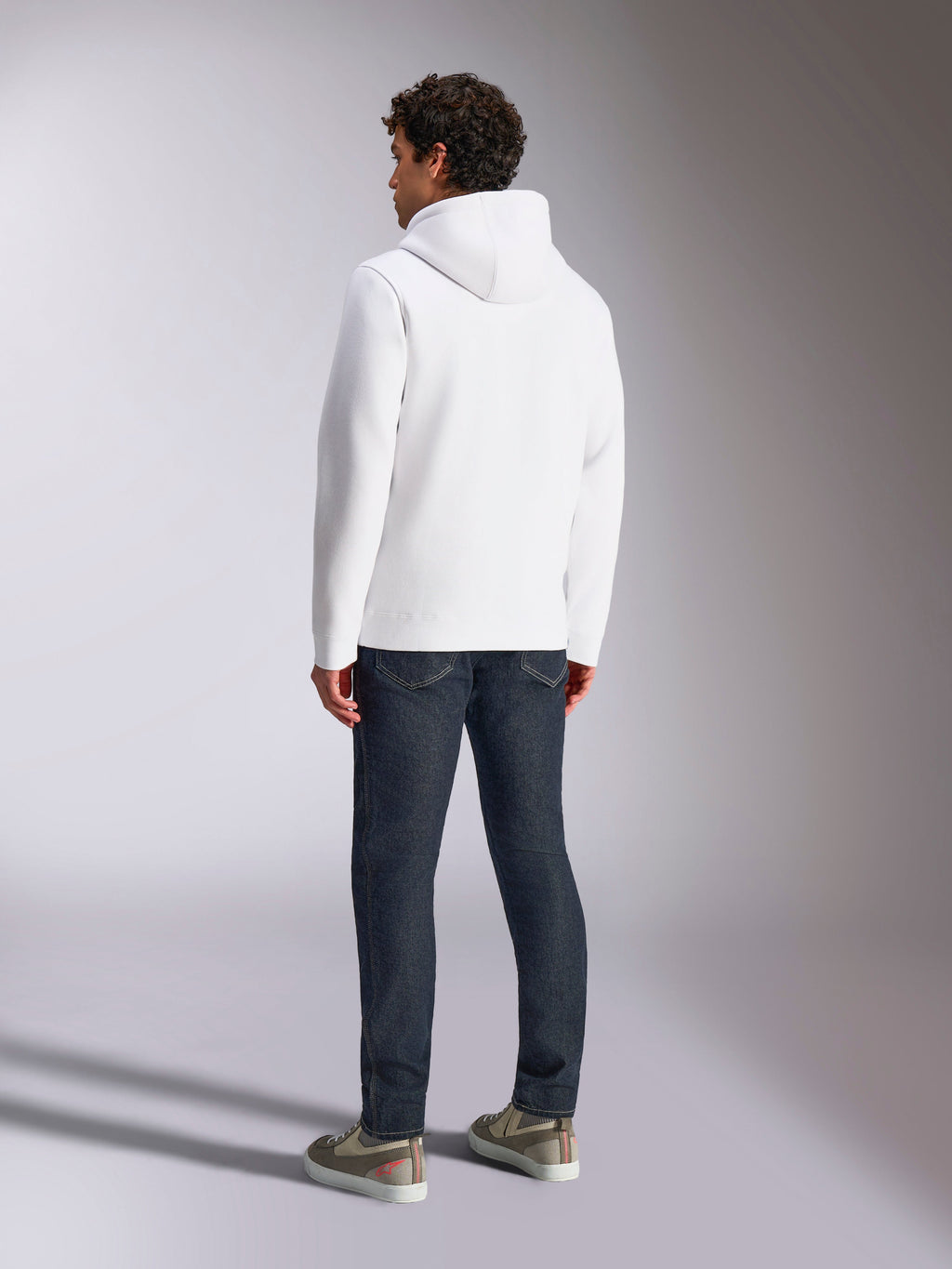 Alpinestars Blaze V3 Hoodie, vista posteriore di un modello maschile che indossa una felpa bianca con cappuccio, abbigliamento sportivo casual abbinato a jeans blu scuro e sneakers grigie, design moderno ispirato al motorsport
