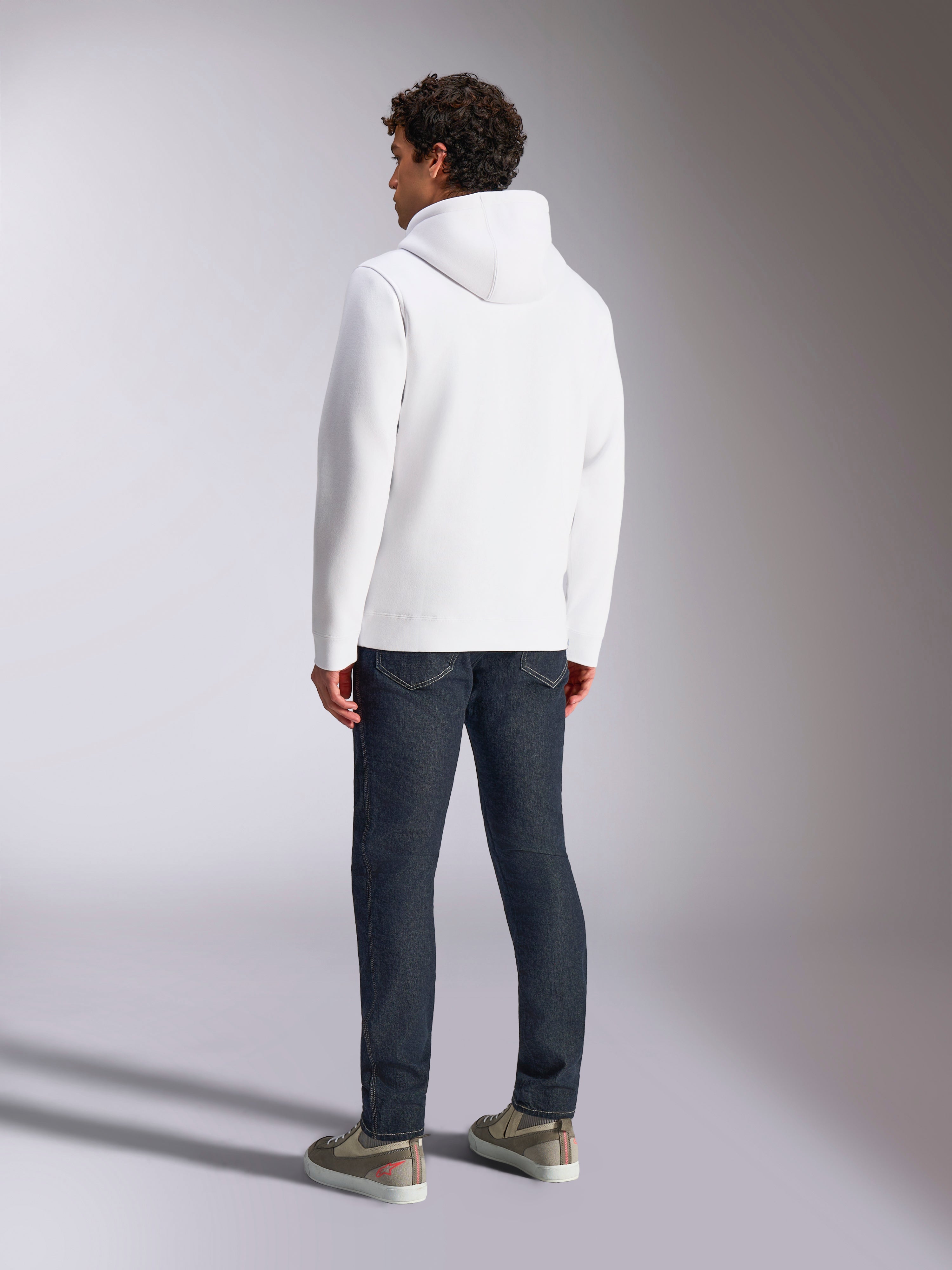 Alpinestars Blaze V3 Hoodie, vista posteriore di un modello maschile che indossa una felpa bianca con cappuccio, abbigliamento sportivo casual abbinato a jeans blu scuro e sneakers grigie, design moderno ispirato al motorsport
