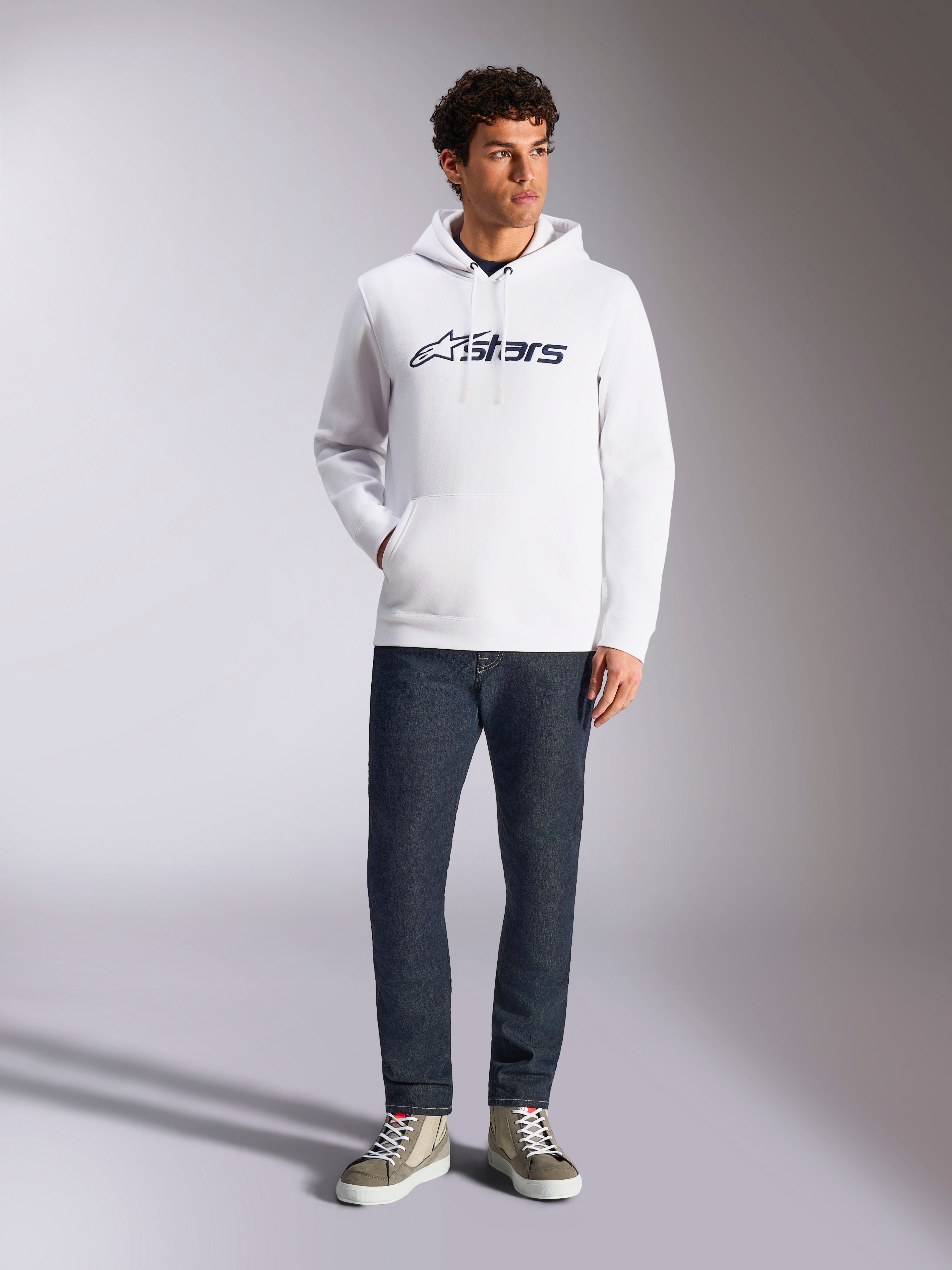 Alpinestars Blaze V3 Hoodie, felpa casual streetwear bianca con logo Alpinestars blu navy sul petto, indossata da un modello con jeans blu scuro e sneakers color sabbia, con cappuccio con coulisse e tasca a marsupio frontale