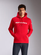 Felpa con cappuccio Alpinestars Linear Wordmark V3, rossa con scritta bianca, streetwear casual, indossata da un modello con le mani nella tasca a marsupio, cappuccio con cordino, jeans in denim scuro, design moderno ispirato al motorsport
