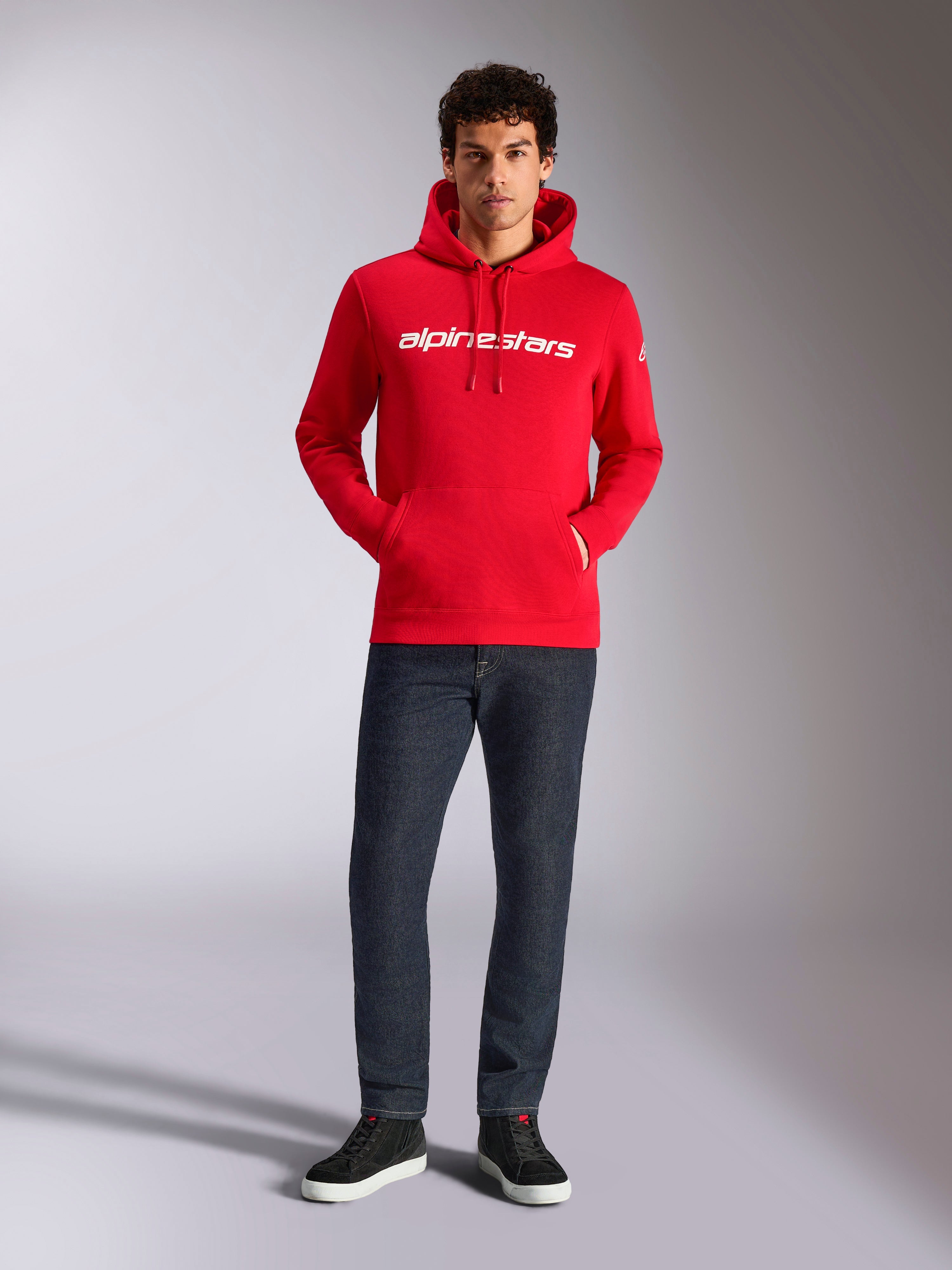 Felpa con cappuccio Alpinestars Linear Wordmark V3, streetwear casual, rossa con scritta lineare bianca sul petto, cappuccio con cordino, tasca a marsupio frontale, indossata con jeans scuri e sneakers nere, design moderno ispirato al motorsport