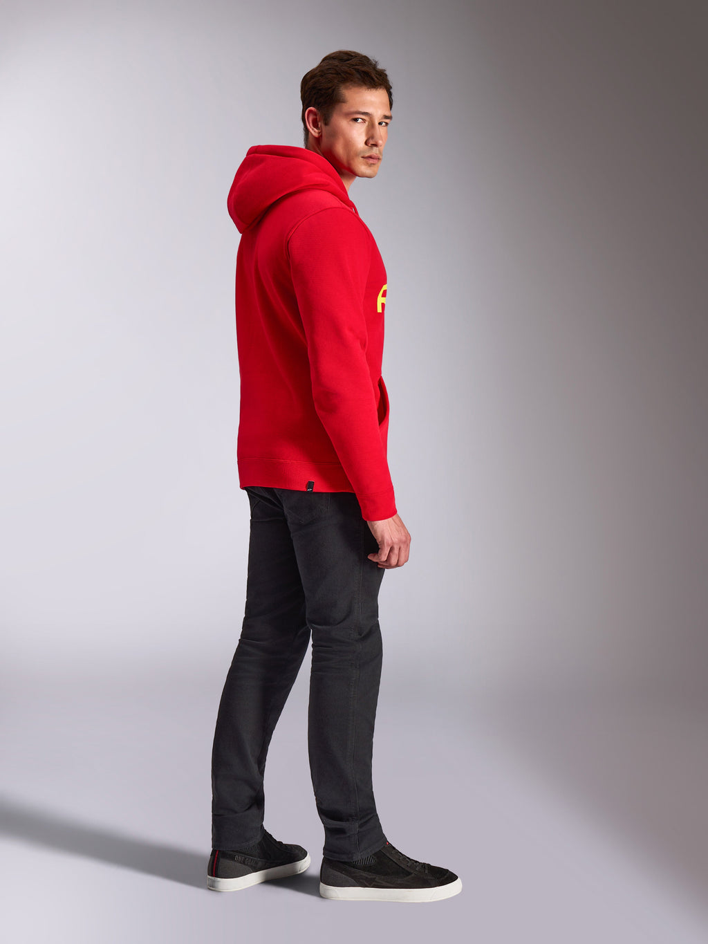 Felpa con cappuccio Alpinestars Linear Wordmark V3, rosso, streetwear casual, modello visto di lato e da dietro che indossa una felpa rossa con marchio giallo, jeans grigio scuro e sneakers nere, scatto lifestyle in studio