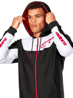 Alpinestars Sessions V3 Hoodie, felpa con cappuccio e zip anteriore, nero, bianco e rosso, indossata da un uomo con il cappuccio alzato, loghi Alpinestars rossi sul petto e sulle maniche, streetwear casual da motorsport