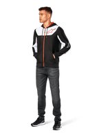Felpa con cappuccio Alpinestars Sessions V3, felpa casual con zip frontale, nero, bianco e rosso, indossata da un modello con jeans scuri e sneakers nere, caratterizzata da pannelli color-block bianchi sul petto e sulle maniche, dettagli della zip e dei cordini rossi, grande logo sul petto, streetwear in pile di poliestere ispirato al motorsport.