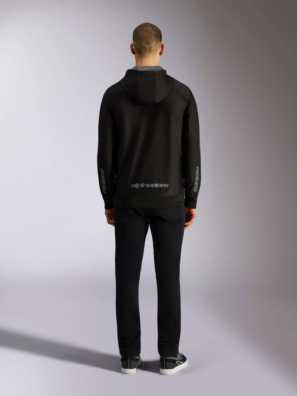Vista posteriore della felpa con cappuccio Alpinestars Sessions V3 in nero e grigio, con scritta Alpinestars grigia sulla parte inferiore della schiena e loghi astars sugli avambracci, indossata da un modello con pantaloni neri, streetwear casual ispirato al motorsport