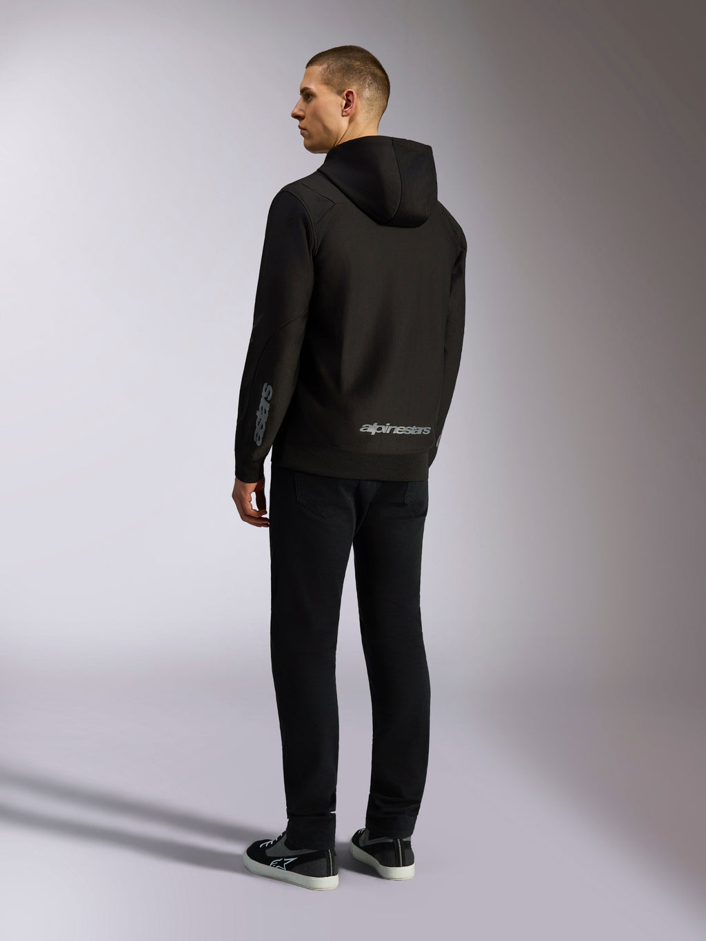 Felpa con cappuccio Alpinestars Sessions V3, streetwear casual nero, vista posteriore su un modello, con scritta Alpinestars grigia sulla parte inferiore della schiena e logo Astars sulla manica, design con cappuccio e zip frontale dallo stile moderno ispirato al motorsport