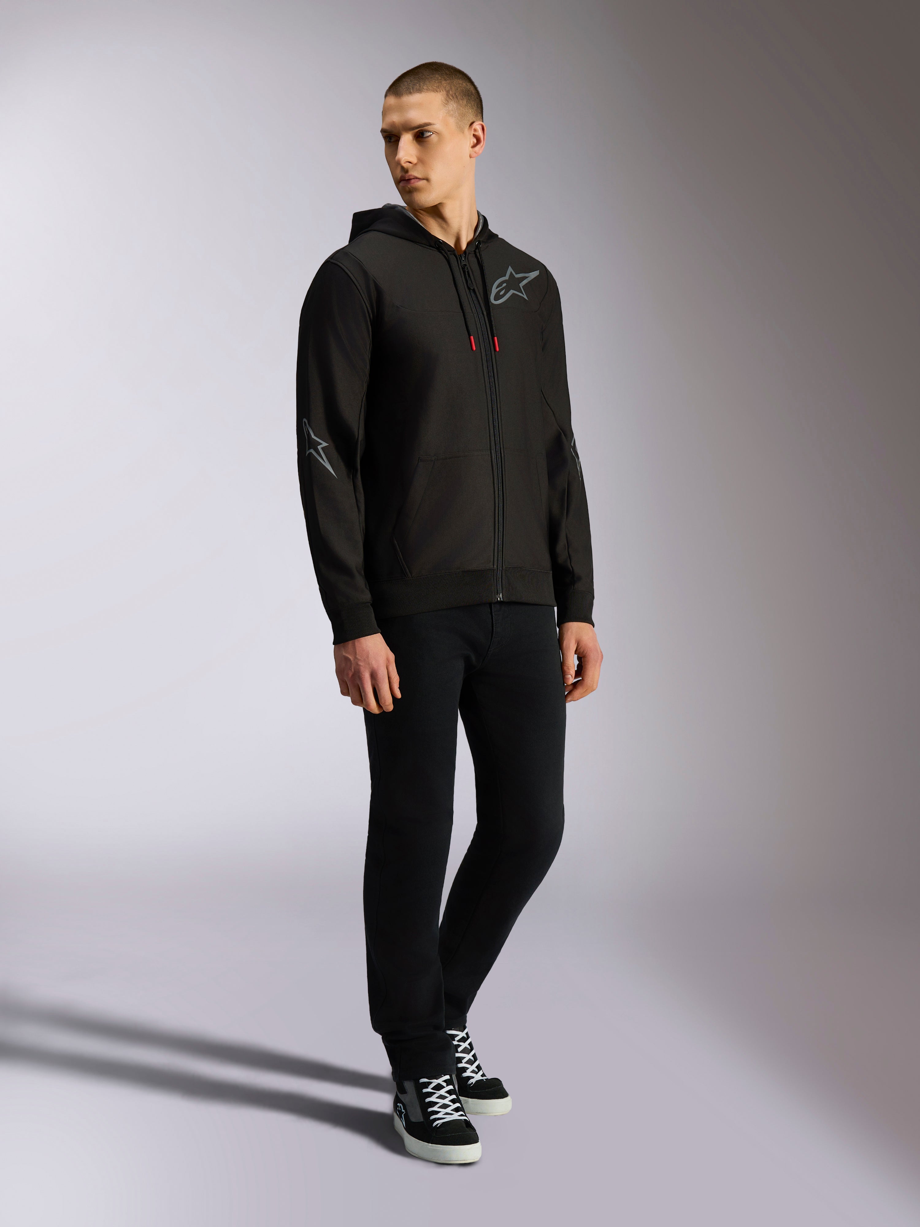 Alpinestars Sessions V3 Hoodie, felpa in pile di poliestere con zip frontale, nera e grigia, loghi grigi su petto e maniche, cappuccio con coulisse e punte dei cordini rosse, vista a figura intera del modello in abbigliamento casual motorsport streetwear