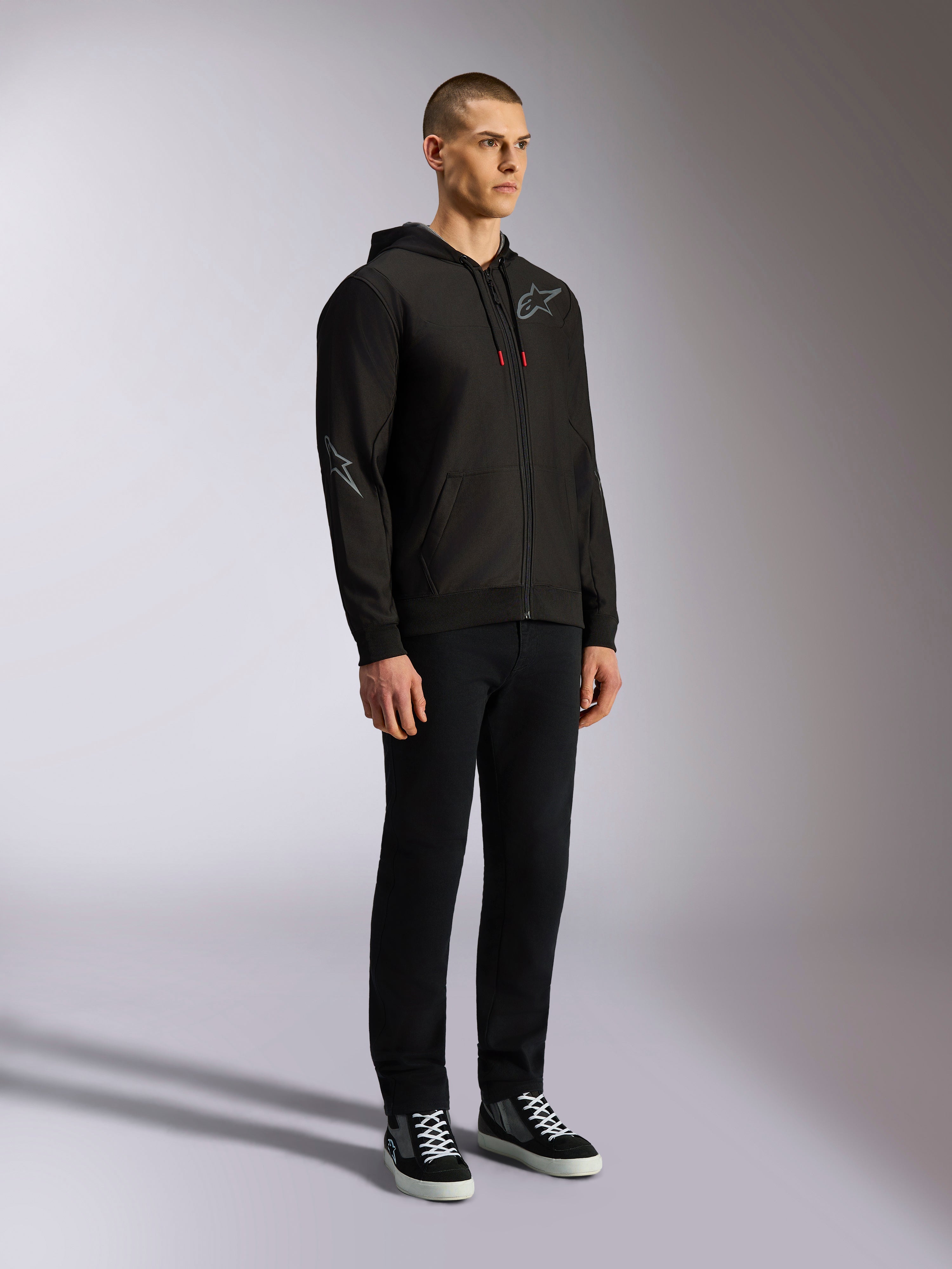 Felpa con cappuccio Alpinestars Sessions V3, streetwear casual da motorsport, felpa nera con zip e logo a stella grigio sul petto e sulla manica, cordini con punte rosse, polsini e orlo a coste, indossata da un modello con pantaloni neri e sneaker.