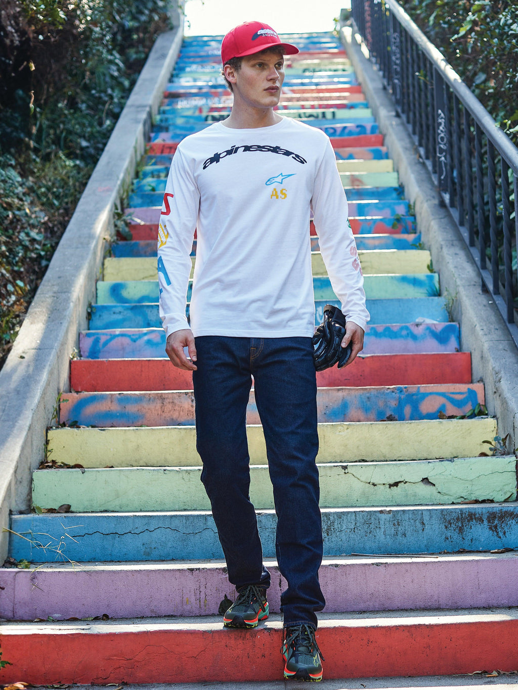 Alpinestars Brundo CSF Maglietta a Maniche Lunghe, t-shirt bianca con logo Alpinestars nero e stampe grafiche colorate, indossata da un modello su scale colorate, abbinata a jeans blu scuro e un cappellino rosso, streetwear casual da motorsport.