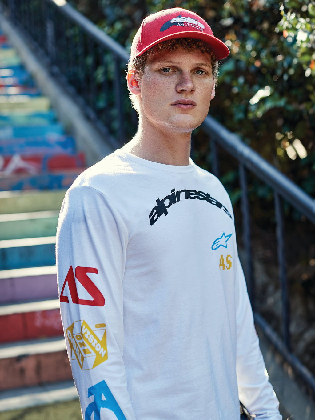 Cappello Alpinestars Profile, rosso, indossato da un giovane uomo, corona strutturata con visiera curva e logo ricamato, abbinato a una T-shirt bianca a maniche lunghe con grafica, streetwear casual ispirato al motorsport