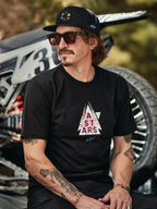 Cappello Alpinestars Gambit Trucker nero con patch logo circolare, indossato con T-shirt Alpinestars Soloist SS nera, scatto lifestyle con un uomo con occhiali da sole e tatuaggi seduto davanti a una moto da cross.