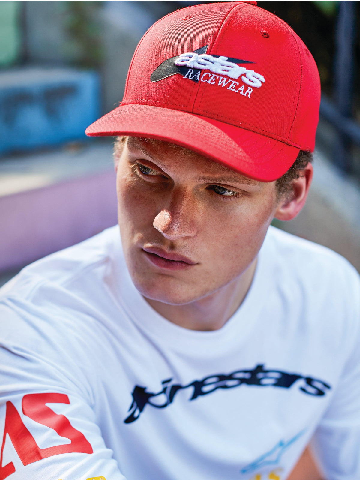 Uomo che indossa il Cappello Alpinestars Profile, snapback rosso con visiera curva, ricamo frontale 'astars RACEWEAR' bianco e nero, abbinato a una t-shirt grafica bianca Alpinestars a maniche lunghe, look casual motorsport lifestyle