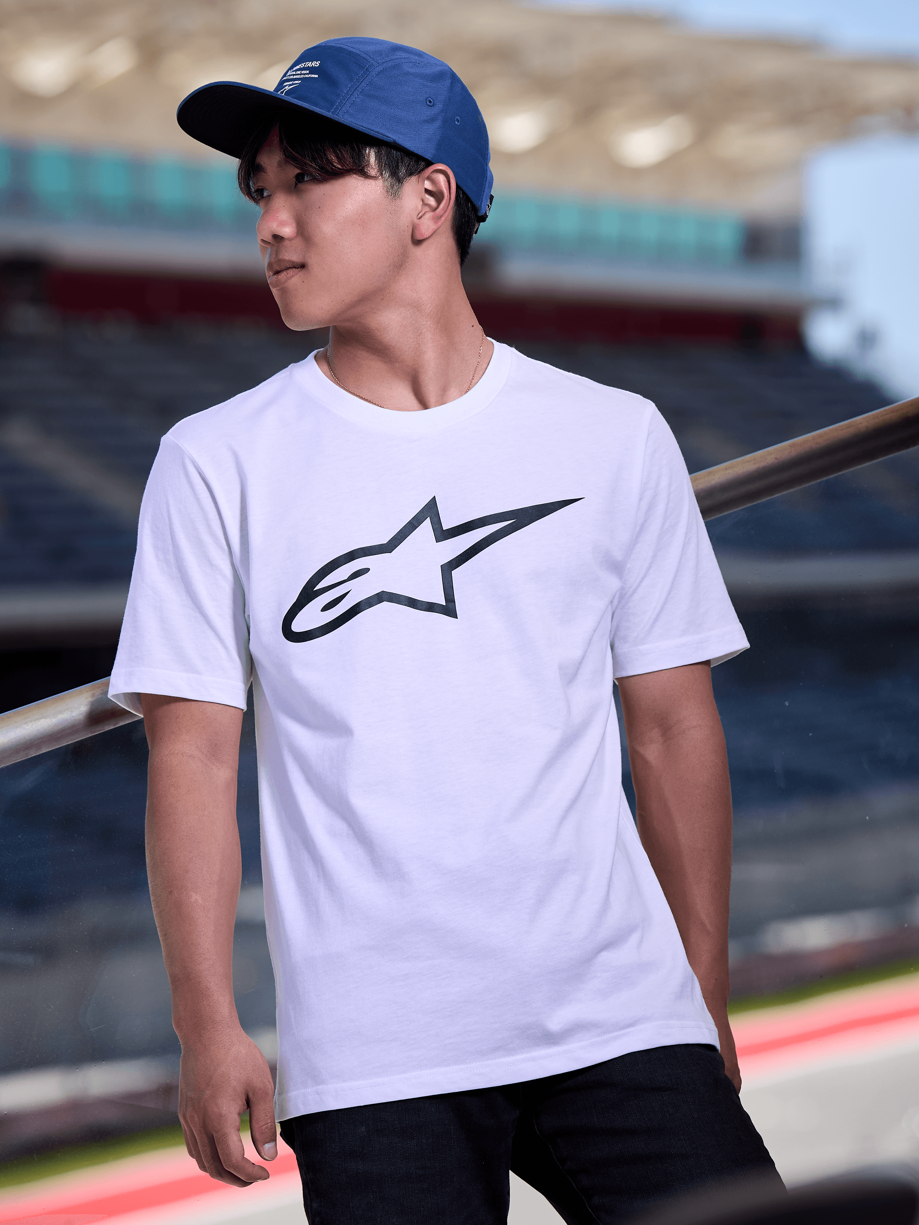 Uomo con cappello Alpinestars Peasy blu, cappello jockey a 5 pannelli con visiera piatta e stampa grafica con logo bianco, abbinato a una T-shirt bianca con logo Alpinestars, streetwear casual ispirato al motorsport