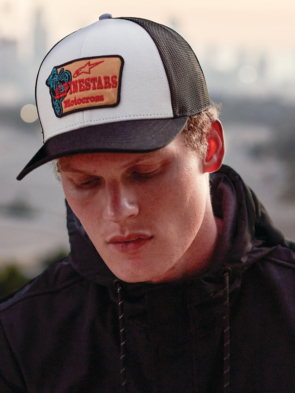 Alpinestars Hardware Trucker Hat in colore Military White, indossato da un uomo con una felpa nera con cappuccio, caratterizzato da un pannello frontale bianco con visiera curva nera e retro in rete nera, patch ricamata color sabbia con marchio Alpinestars Motocross rosso, stile casual lifestyle motorsport streetwear