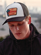 Alpinestars Hardware Trucker Hat in colore Military White, indossato da un uomo con una felpa nera con cappuccio, caratterizzato da un pannello frontale bianco con visiera curva nera e retro in rete nera, patch ricamata color sabbia con marchio Alpinestars Motocross rosso, stile casual lifestyle motorsport streetwear