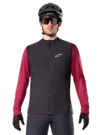 Gilet termico Alpinestars A-Dura, gilet da ciclismo isolato nero, indossato da un modello con maglia bordeaux a maniche lunghe, casco da MTB nero e occhiali da sole, con logo bianco sul petto, protezione termica leggera per mountain bike