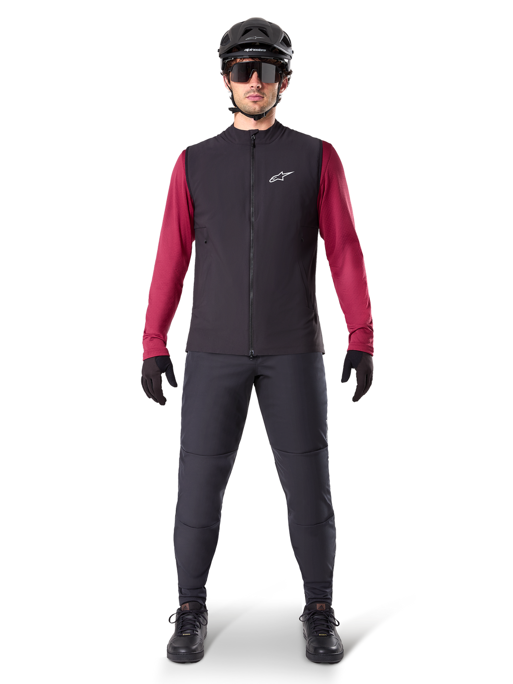 Gilet Termico Alpinestars A-Dura, gilet da ciclismo nero indossato sopra una maglia a maniche lunghe bordeaux, abbinato a pantaloni da MTB grigio scuro, casco e occhiali da sole neri, abbigliamento tecnico da mountain bike per climi freddi