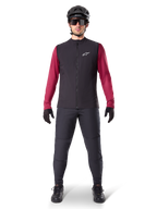 Gilet Termico Alpinestars A-Dura, gilet da ciclismo nero indossato sopra una maglia a maniche lunghe bordeaux, abbinato a pantaloni da MTB grigio scuro, casco e occhiali da sole neri, abbigliamento tecnico da mountain bike per climi freddi