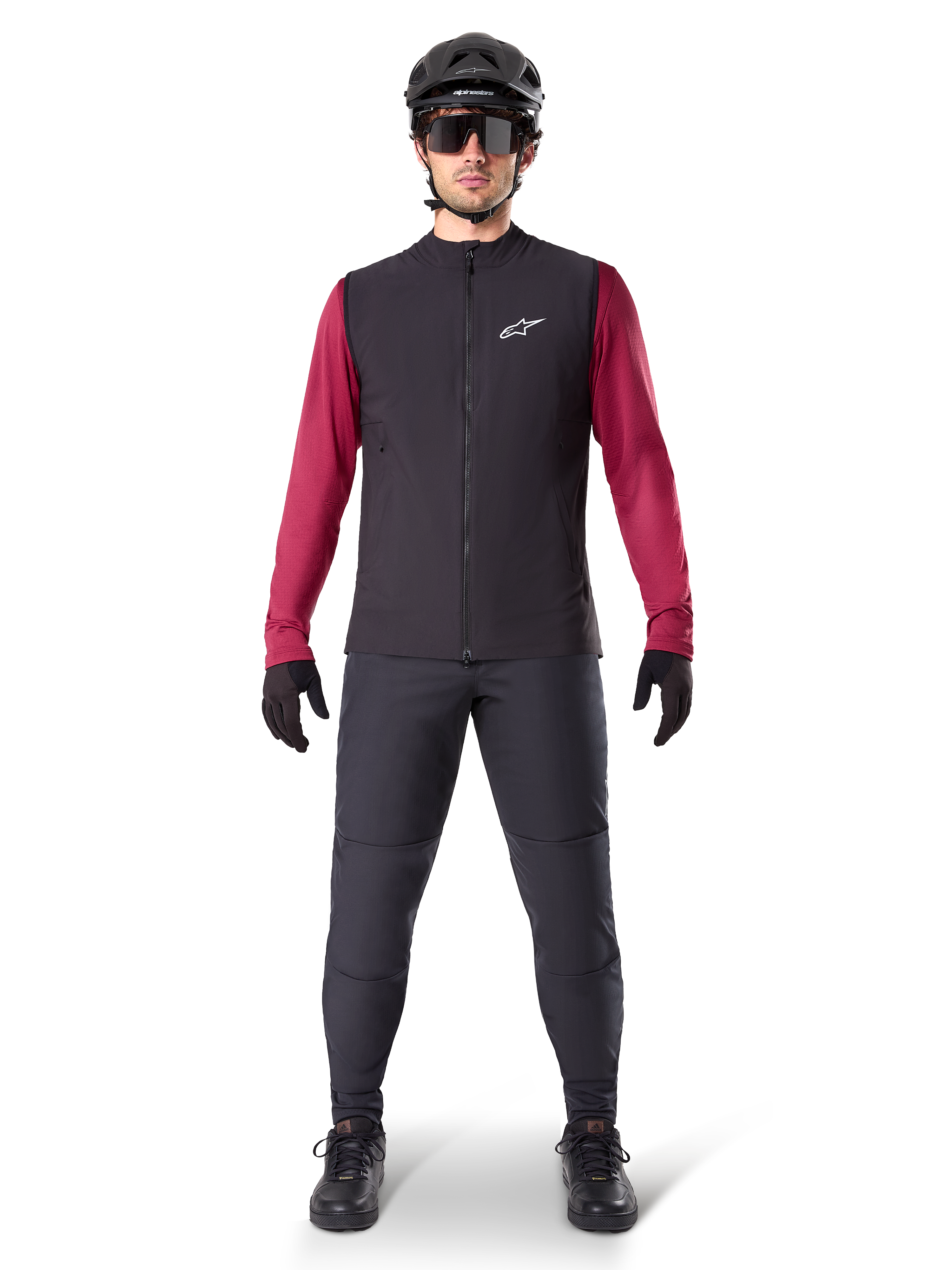 Gilet Termico Alpinestars A-Dura, gilet da ciclismo nero indossato sopra una maglia a maniche lunghe bordeaux, abbinato a pantaloni da MTB grigio scuro, casco e occhiali da sole neri, abbigliamento tecnico da mountain bike per climi freddi