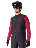 Alpinestars A-Dura Thermal Vest, gilet termico nero da ciclismo, indossato sopra una maglia bordeaux a maniche lunghe, con logo bianco sul petto, abbinato a un casco da MTB nero e occhiali da sole, strato isolante performante per mountain bike