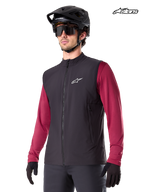 Alpinestars Gilet Termico A-Dura, gilet termico da mountain bike da uomo in nero, indossato da un modello con casco e maglia a maniche lunghe rossa, isolamento per la guida MTB in climi freddi