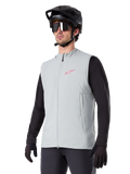 Alpinestars A-Dura Thermal Vest, grigio titanio, gilet termico da ciclismo, indossato sopra una maglia nera a maniche lunghe con casco da mountain bike e guanti neri, con logo rosa sul petto e tasche laterali verticali con zip