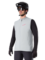 Alpinestars A-Dura Thermal Vest, grigio titanio, gilet termico da ciclismo, indossato sopra una maglia nera a maniche lunghe con casco da mountain bike e guanti neri, con logo rosa sul petto e tasche laterali verticali con zip
