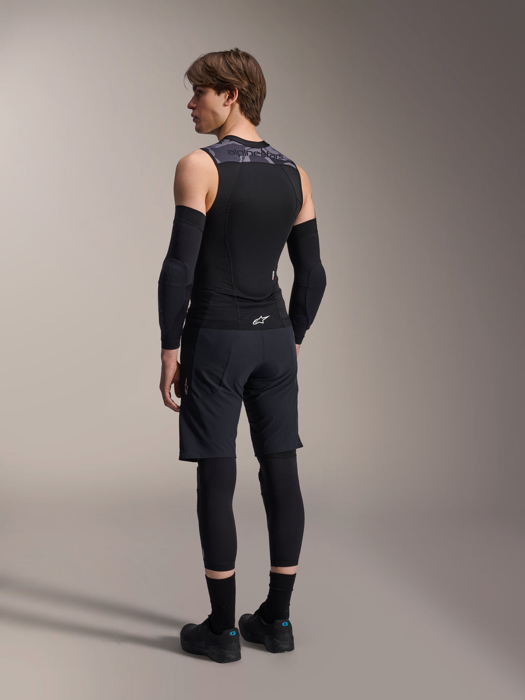 Modello maschile visto di spalle che indossa il gilet protettivo Alpinestars A-Motion Plasma, Black Camo, design in mesh senza maniche con paraschiena integrato, motivo mimetico grigio sulle spalle, abbinato a gomitiere nere e pantaloncini da mountain bike, abbigliamento tecnico da ciclismo