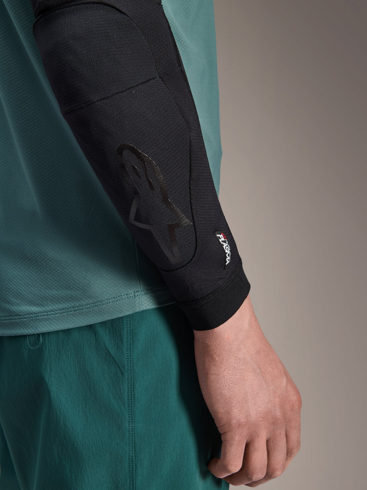 Alpinestars A-Motion Plasma Elbow Sleeve, gomitiera nera per MTB, vista ravvicinata indossata con maglia color verde acqua, logo Astars tono su tono e marchio Nucleon Plasma, design in mesh a profilo sottile per ciclismo fuoristrada