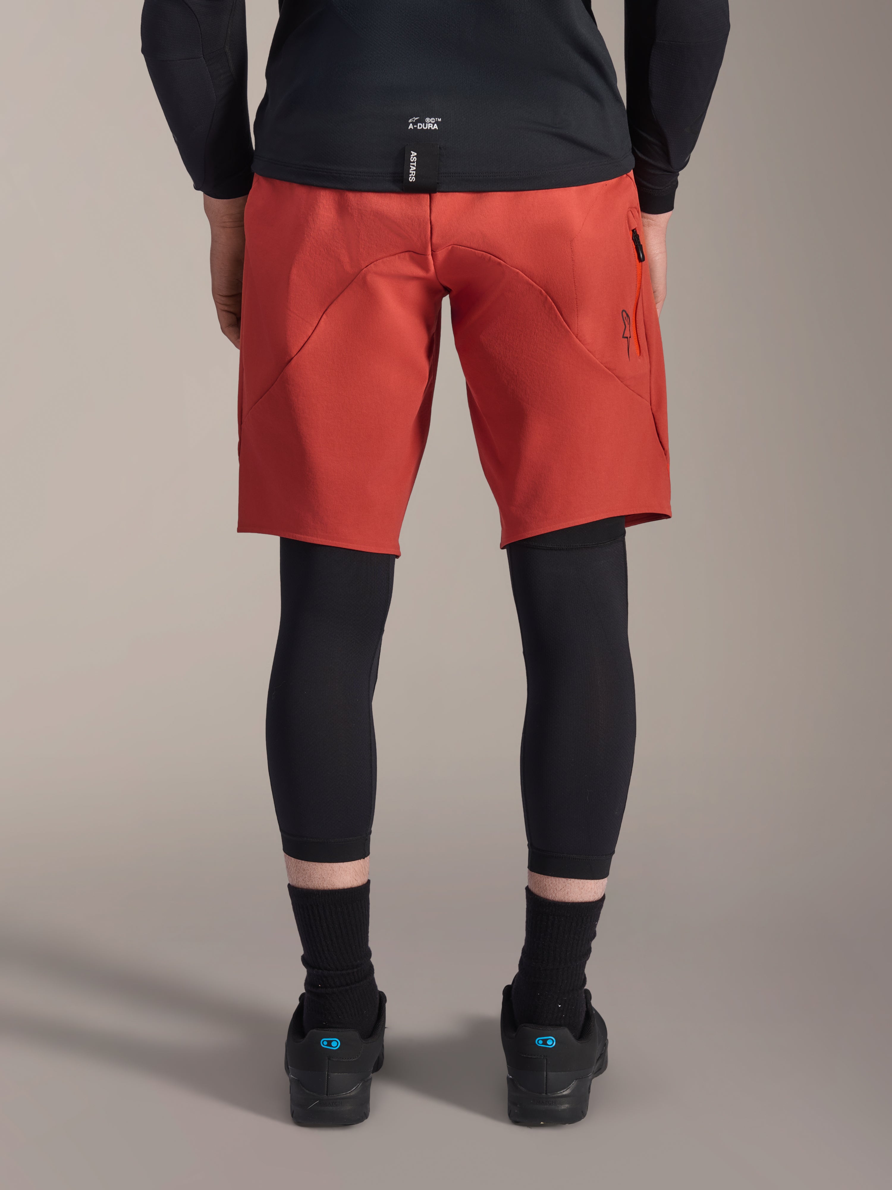 Alpinestars A-Motion Plasma Knee Sleeve, protezioni per ginocchia da MTB nere, vista posteriore indossate sotto pantaloncini rossi da mountain bike con maglia nera e scarpe tecniche da ciclismo, attrezzatura protettiva per mountain bike