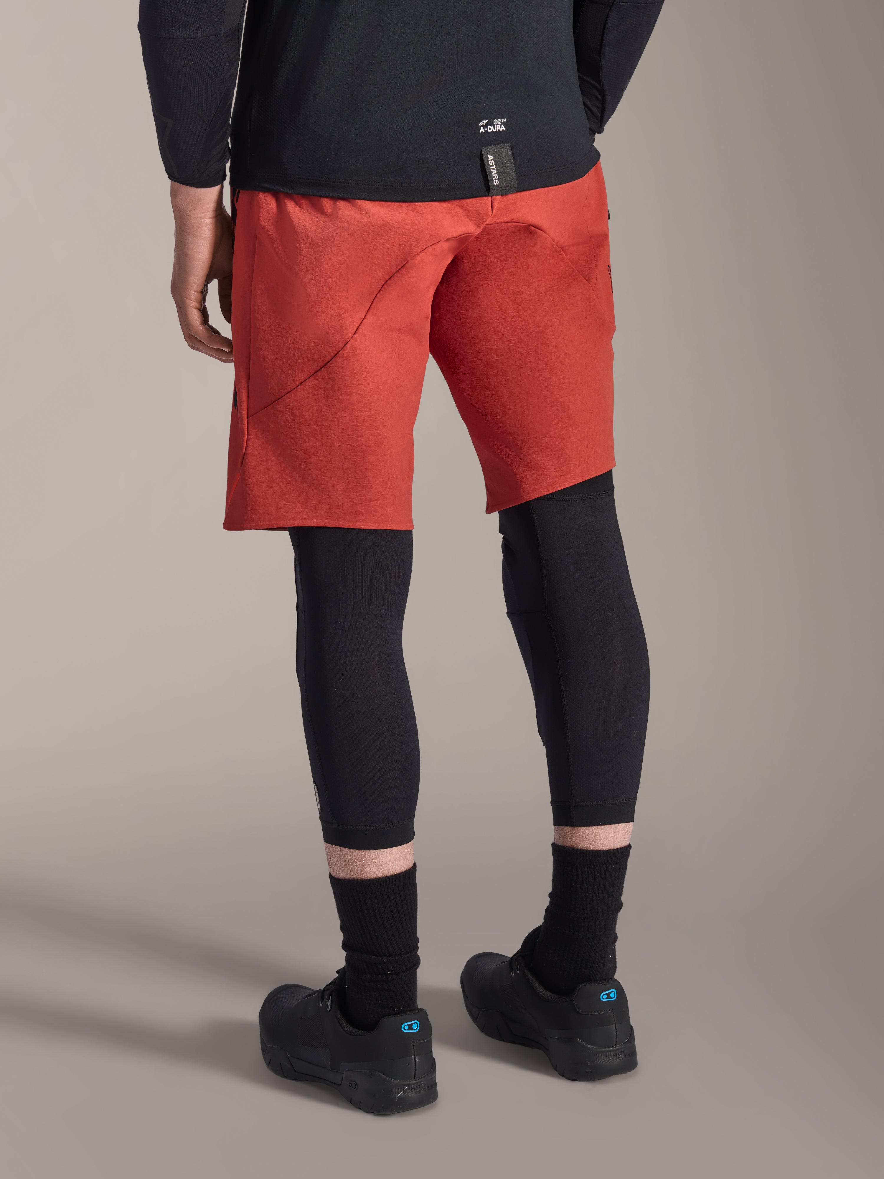 Alpinestars A-Motion Plasma Knee Sleeve, ginocchiera MTB nera, indossata con pantaloncini da MTB rossi e scarpe da ciclismo nere, vista posteriore con design a infilare a basso profilo per ciclismo fuoristrada
