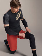 Ginocchiera Alpinestars A-Motion Plasma, protezione MTB, nero, indossata da un rider in posizione inginocchiata, abbinata a una t-shirt nera e grigia con logo oro, pantaloncini rossi e gomitiere nere, equipaggiamento protettivo professionale per mountain bike