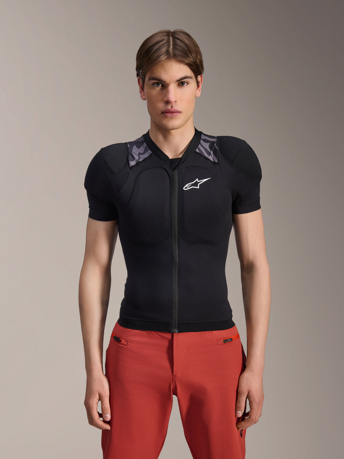Modello che indossa la giacca protettiva Alpinestars A-Motion Plasma - Short Sleeve, nera con imbottitura mimetica grigia sulle spalle, logo Alpinestars bianco sul petto, abbinata a pantaloni rossi, protezione per il corpo MTB slim-fit con zip frontale