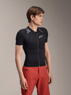 Modello che indossa la giacca protettiva Alpinestars A-Motion Plasma a maniche corte, colore Black Camo con pannelli mimetici grigi sulle spalle, dotata di protezioni integrate per spalle e torace, logo a stella bianco sul petto, tessuto a rete traspirante, abbinata a pantaloni rossi