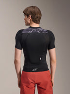 Giacca protettiva Alpinestars A-Motion Plasma - Maniche Corte, nero con dettagli mimetici grigi sulla parte superiore della schiena, vista posteriore con paraschiena integrato e logo Astars bianco sull'orlo, indossata con pantaloncini rossi, abbigliamento protettivo tecnico per mountain bike