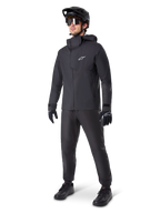 Mountain biker uomo con pantaloni impermeabili Alpinestars A-Dura neri, abbinati a giacca impermeabile nera con cappuccio coordinata, casco da MTB nero con visiera, occhiali da ciclismo scuri e guanti neri, completo professionale per trail riding in ogni condizione
