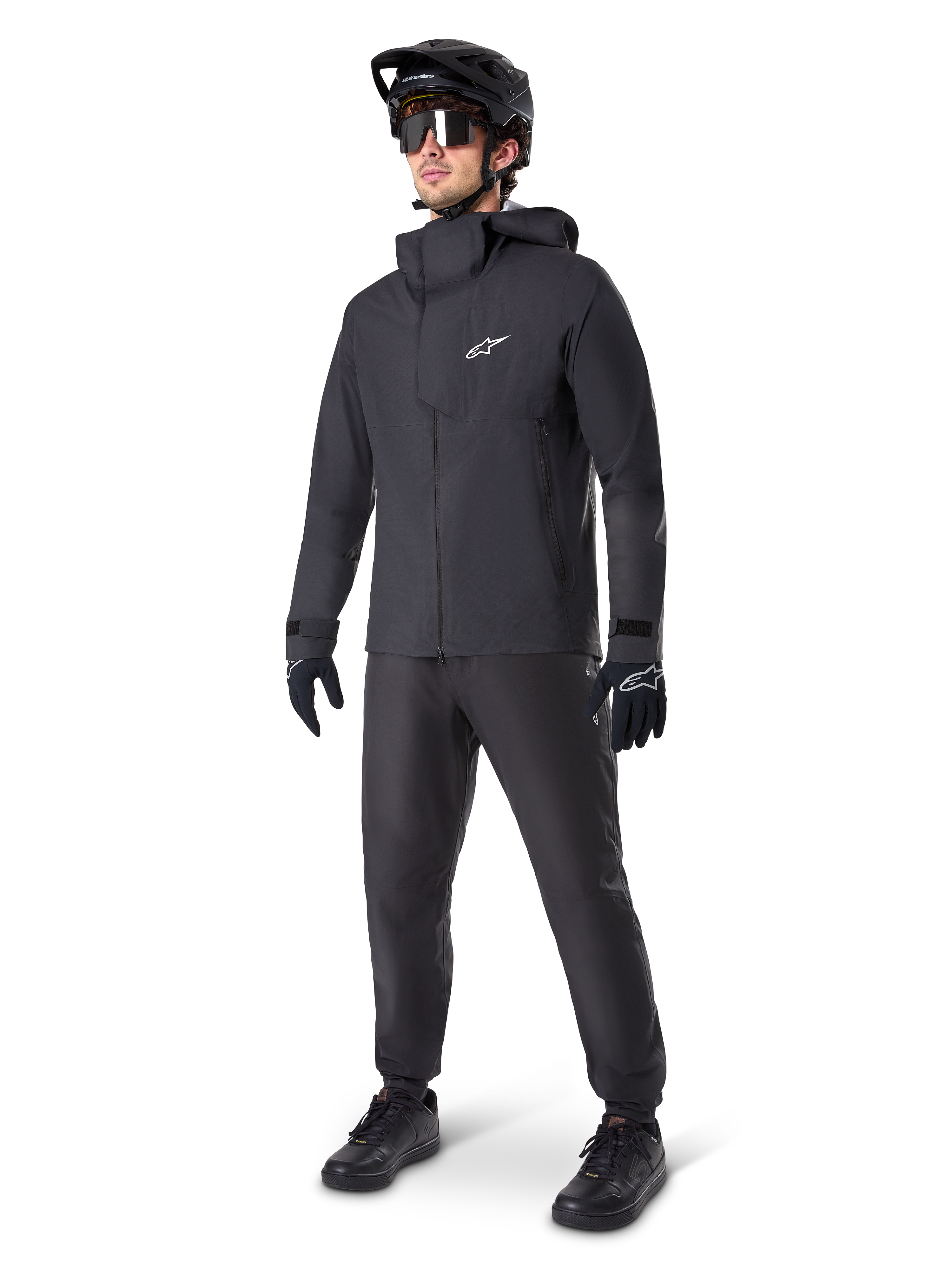 Mountain biker uomo con pantaloni impermeabili Alpinestars A-Dura neri, abbinati a giacca impermeabile nera con cappuccio coordinata, casco da MTB nero con visiera, occhiali da ciclismo scuri e guanti neri, completo professionale per trail riding in ogni condizione
