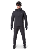 Pantaloni impermeabili Alpinestars A-Dura, pantaloni da MTB neri, indossati come parte di un set completo di abbigliamento da mountain bike total black che include giacca tecnica impermeabile, casco nero, occhiali da sole e guanti, progettati per la guida fuoristrada in condizioni di pioggia.