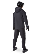 Pantaloni impermeabili Alpinestars A-Dura, pantaloni da MTB, nero, visti dal retro su un ciclista con abbigliamento da mountain bike nero completo di giacca con cappuccio e casco, abbigliamento impermeabile resistente per sentieri fangosi